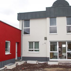 Längliches Hauptgebäude mit grauem Flachdach, weißen Fensterrahmen, großzügigem Eingangsbereich mit viel Glas und grau abgesetztem Sockel. Links und rechts schließen sich kubusfärmige Gebäudeteile an, die in einem tiefen Rot gehalten sind.