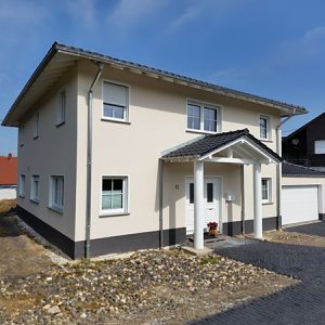 Zweistöckiges, freistehendes Einfamilienhaus mit einem überdachten eingangsbereich mit Satteldach und zwei Säulen, die das Dach abstützen. Graues Dach, weiße Fensterrahmen. Fassade und Säulen in einem hellen Cremeton gehalten.