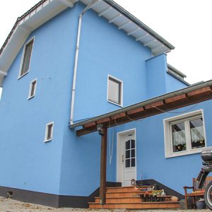 Architektonisch auffällig gestaltetes, freistehendes Einfamilienhaus mit einem Gebäudeteil, der über drei Etagen geht und nach hinten versetzt weitergeführt wird. Rechts schließt sich ein eingeschossiger Gebäudeteil mit Haupteingang und Veranda-Überdachung an. Weiße Fensterrahmen und Dachgaube harmonieren mit einem hellblauen Anstrich.