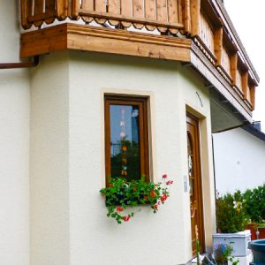Klassisches Landhaus mit Erker, in dem sich Eingangstür und Fenster befinden und über dem ein Holzbalkon liegt. Fassade in einem hellen Gelb / Cremeton gehalten, setzt einen angenehmen und natürlichen Kontrast zum warmen Holz.