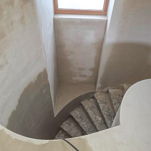 Blick von oben auf eine betongegossene Wendeltreppe. Die Treppe führt an einer Hauswand mit Erker entlang. Der innenliegende Teil der Treppe wurde ebenfalls an einer abgerundeten Wand entlanggeführt. Die Wand wird am oberen Ende der Treppe als Balustrade weitergeführt, um den offenen Treppenraum abzugrenzen.