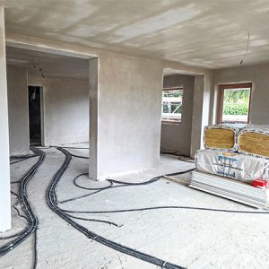 Rohbau von Wohnräumen, in denen Kabel und Leitungen auf dem Boden leigen und Dämmwolle an der Seite steht. Offen gestalteter Raumtrenner trennt einen potenziellen Wohnbereich von Küche und Essbereich.