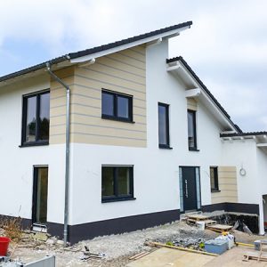 Modernes Haus mit versetztem Pultdach, das an einer Seite über der Garage bis zum Boden gezogen ist. Graue Fensterrahmen und Türen in unterschiedlichen Grüßen sorgen für einen unruhigen und trotzdem symetrischen Look, der durch die Fassadengestaltung in weiß ausgeglichen wird. Ein Teil der Fasse ist hervorgehoben durch Farbgestaltung in Creme mit grauen Zierstreifen, so dass eine Art Holzoptik entsteht.