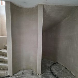 Treppenaufgang einer gegossenen Wendeltreppe im Keller. Die Rundung des Treppenaufgangs findet sich in der Kellerwand neben der Treppe wieder, die halbförmig abgerundet ist. Daneben befindet sich eine Nische. Alles ist noch im Rohbau befindlich und unverputzt.