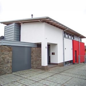 Modernes Einfamilienhaus mit Flachdach. Links ist eine Garage angebaut, die mit Naturstein verkleidet ist. Das Haus selbst hat einen weißen Hauptteil, einen grau verkleideten kleineren Anbau nach hinten raus und rechts einen rot hervorgehobenen Teil. Alles wirkt harmonisch.