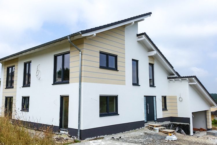 Modernes Haus mit versetztem Pultdach, das an einer Seite über der Garage bis zum Boden gezogen ist. Graue Fensterrahmen und Türen in unterschiedlichen Grüßen sorgen für einen unruhigen und trotzdem symetrischen Look, der durch die Fassadengestaltung in weiß ausgeglichen wird. Ein Teil der Fasse ist hervorgehoben durch Farbgestaltung in Creme mit grauen Zierstreifen, so dass eine Art Holzoptik entsteht.