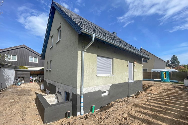 Freistehendes Einfamilienhaus, bei dem das Fundament freigelegt und trockengelegt wurde. Die Fassade in einem grau-grün ist gereinigt und für das Streichen vorbereitet worden.