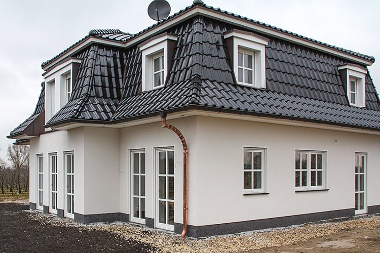 Villenähnliches Einfamilienhaus mit grauem Mansardendach und Schopf. Im senkrecht stehenden Dachteil sind Fenster eingelassen. Die Fassade ist in einem hellen Cremeton gestrichen, der mit Fensterrahmen und dunkelgrauem Dach harmoniert.